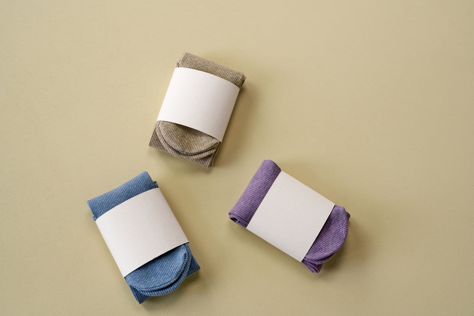 Three rolled pairs of ribbed socks in tan, blue, and purple, each wrapped with a paper band, on a flat lay